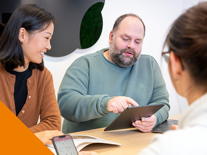 Zwei Frauen und ein MaNn sitzen um einen Tisch. Sie arbeiten am Laptop uind Handy.