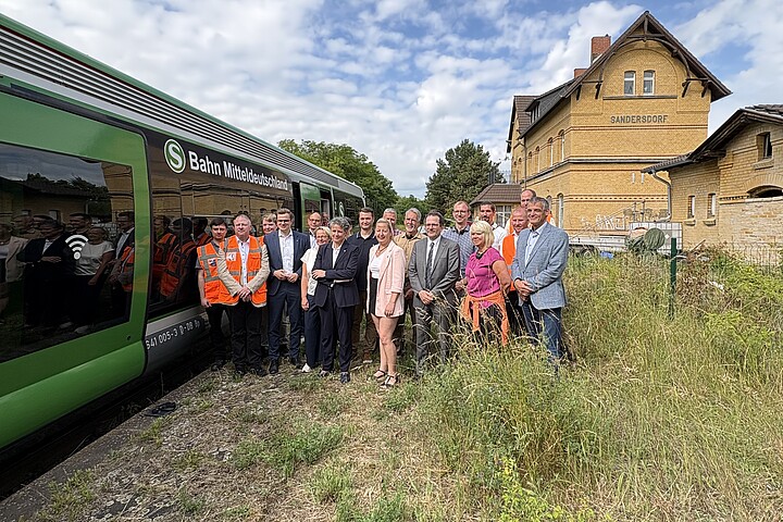 Gruppenbild, unter anderem mit Ministerin Hüskens und Staatssekretär Haller, an einem zugewachsenen Bahnsteig. Links neben der Gruppe steht ein grün-weißer Zug. Rechts ist ein Bahnhofsgebäude mit der Aufschrift Sandersdorf zu sehen.