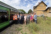 Gruppenbild, unter anderem mit Ministerin Hüskens und Staatssekretär Haller, an einem zugewachsenen Bahnsteig. Links neben der Gruppe steht ein grün-weißer Zug. Rechts ist ein Bahnhofsgebäude mit der Aufschrift Sandersdorf zu sehen.