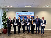 Drei Frauen und fünf Männer in dunklen Anzügen stehen in einem Raum vor einem Bildschirm. Auf dem Bildschirm ist das Modell eines orange-weißen Zuges zu sehen. Die Personen halten Vertragsunterlagen in den Händen.