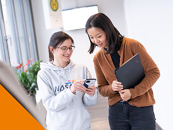 Wine Frau mit einem grauen Pullover auf den das Logo der NASA GmbH gedruckt ist, zeigt einer anderen Frau etwas auf einem Handy