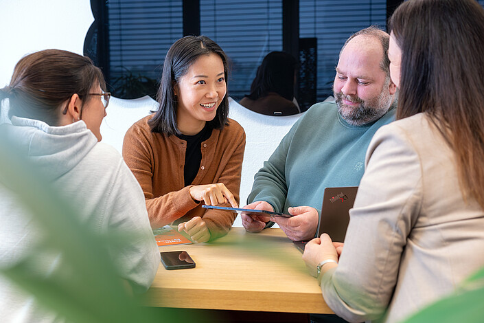 Drei Frauen und ein Mann sitzen um einen Tisch. Sie arbeiten am Laptop und Handy.