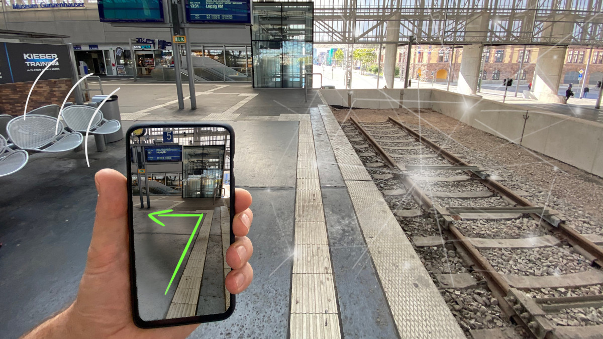 Eine Person hält in einem Bahnhof ein Smartphone in der Hand. Auf dem Bildschirm ist ein Live-Foto der Handykamera zu sehen, auf dem ein grüner Pfeil den Weg durch das Gebäude weist.